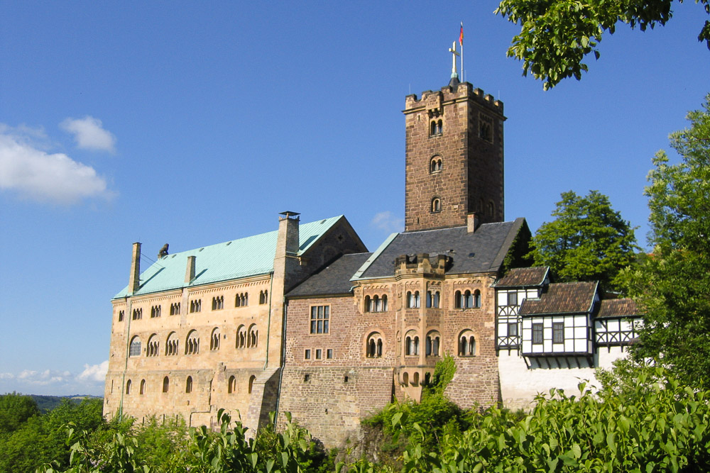 The west side of Wartburg Castle is shown here along with the great hall, keep and Elisabethgang (St. Elisabeth Passage). The stone walls of the buildings are plastered towards the front part of the view; further to the south are rough stone walls, seen towering over rugged cliffs.