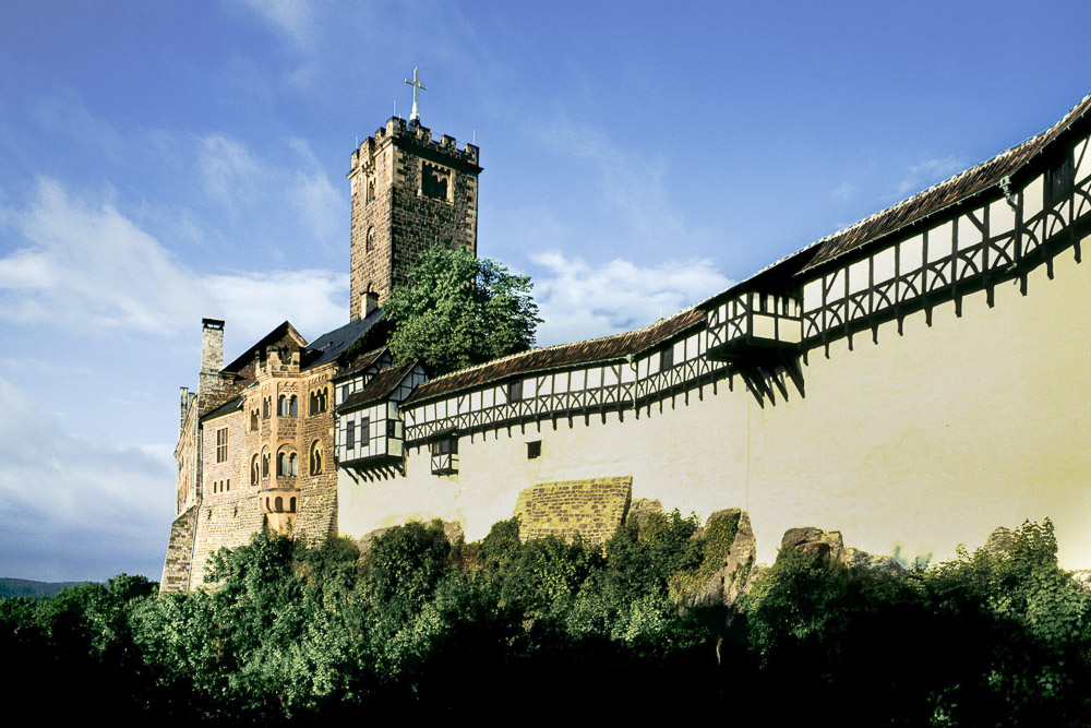 The west side of Wartburg Castle is shown here along with the great hall, keep and Elisabethgang (St. Elisabeth Passage). The stone walls of the buildings are plastered towards the front part of the view; further to the south are rough stone walls, seen towering over rugged cliffs.